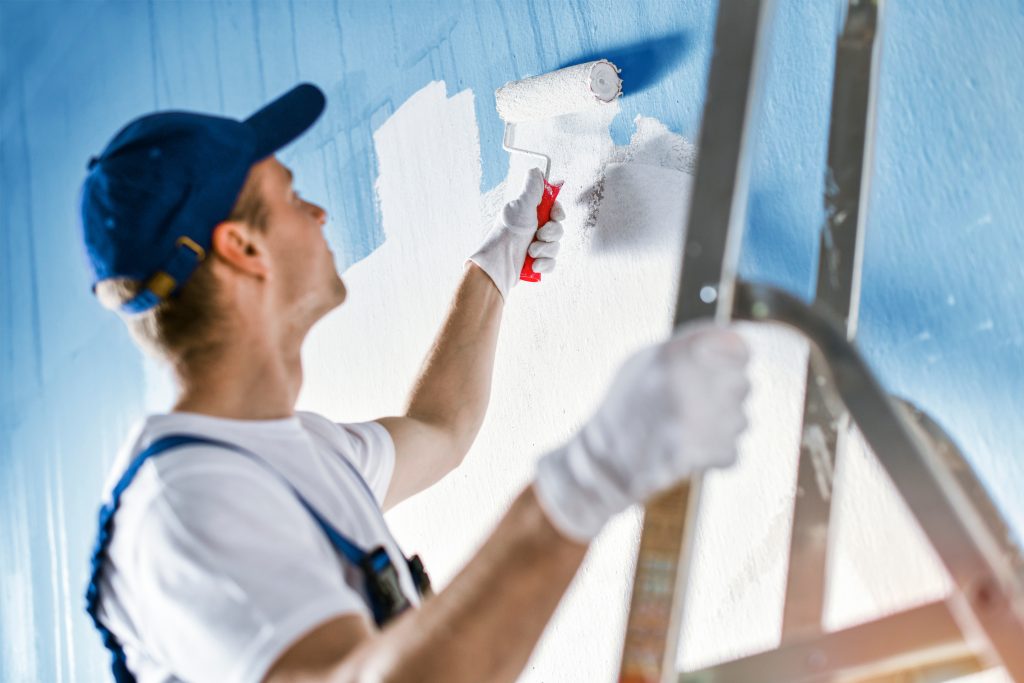 Painter painting a wall with paint roller. Builder worker painting blue surface with white color Fassaden-Sanierung vom Profi